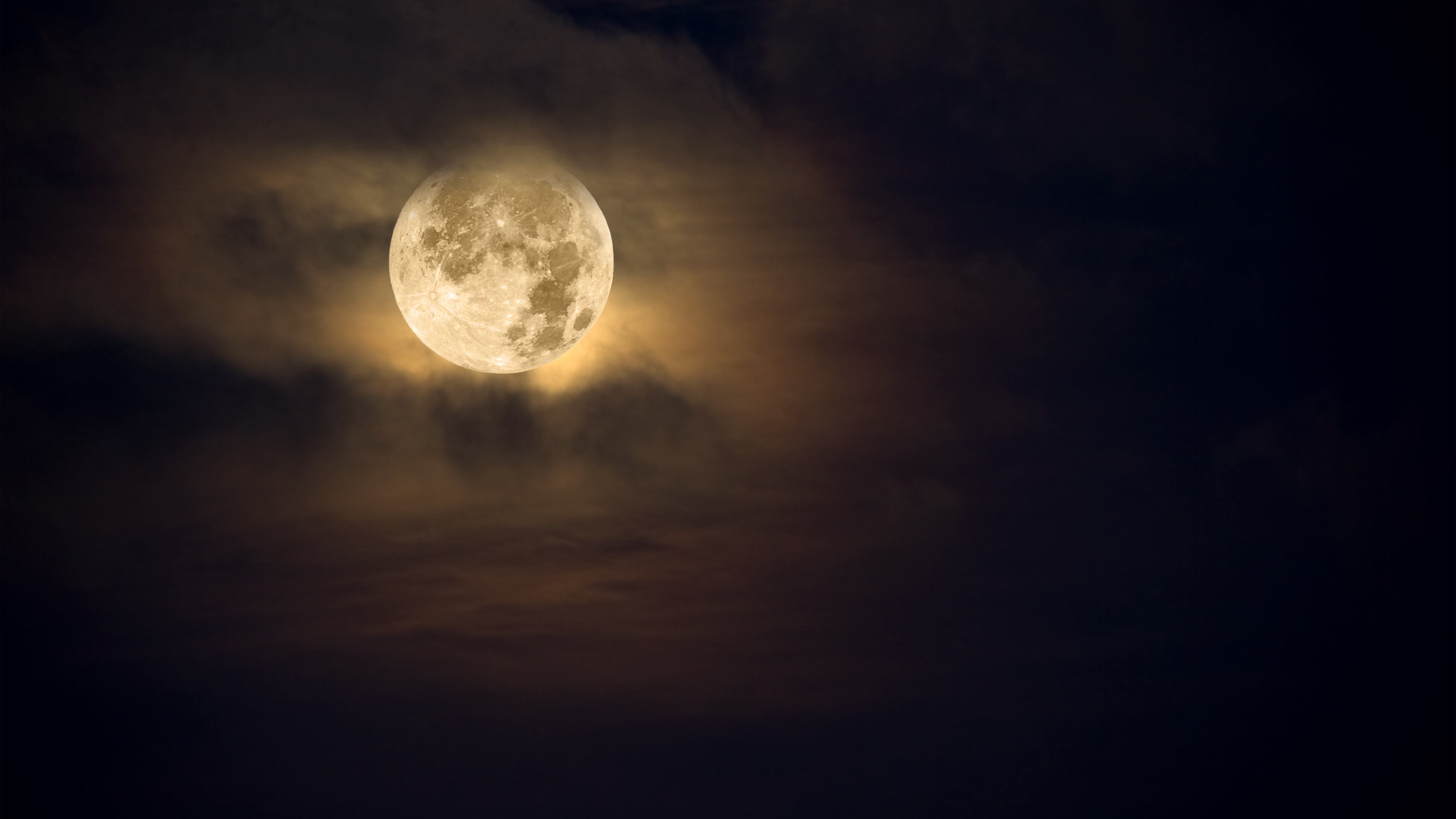A dark night brings a bright, amber moon alive with puffy hazy clouds.